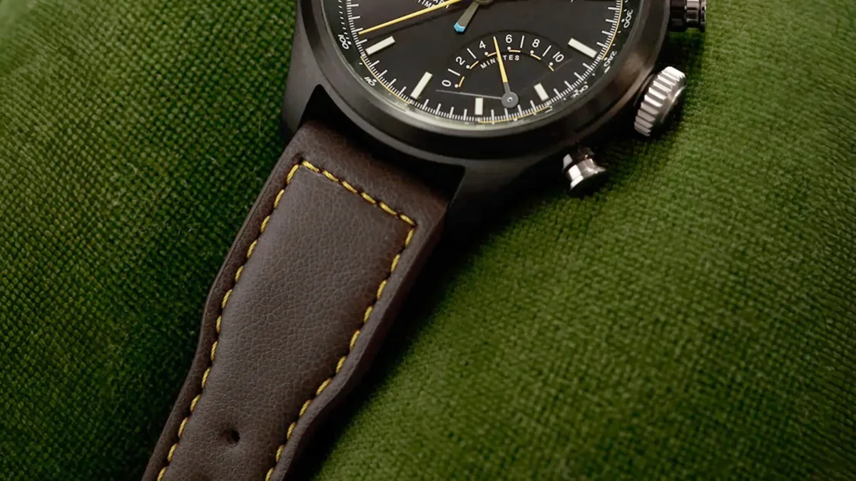 Close-up of a black watch face with yellow and blue hands and a brown leather strap with yellow stitching on green fabric.