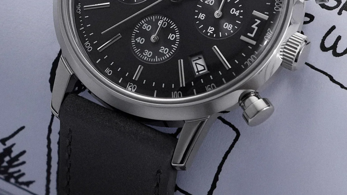 Close-up of a black chronograph watch face with silver markers, date window showing 14, and black leather strap.