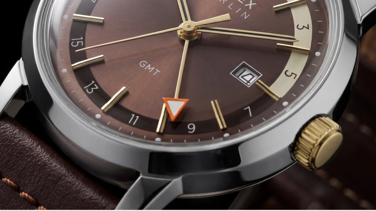 Luxury wristwatch with brown dial, gold hands, orange-tipped GMT hand, date window at 3 o’clock, and a brown leather strap.