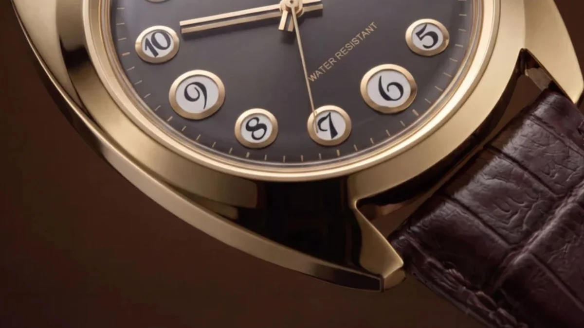 Close-up of a gold watch with a dark dial, round white number markers, and a brown leather strap.